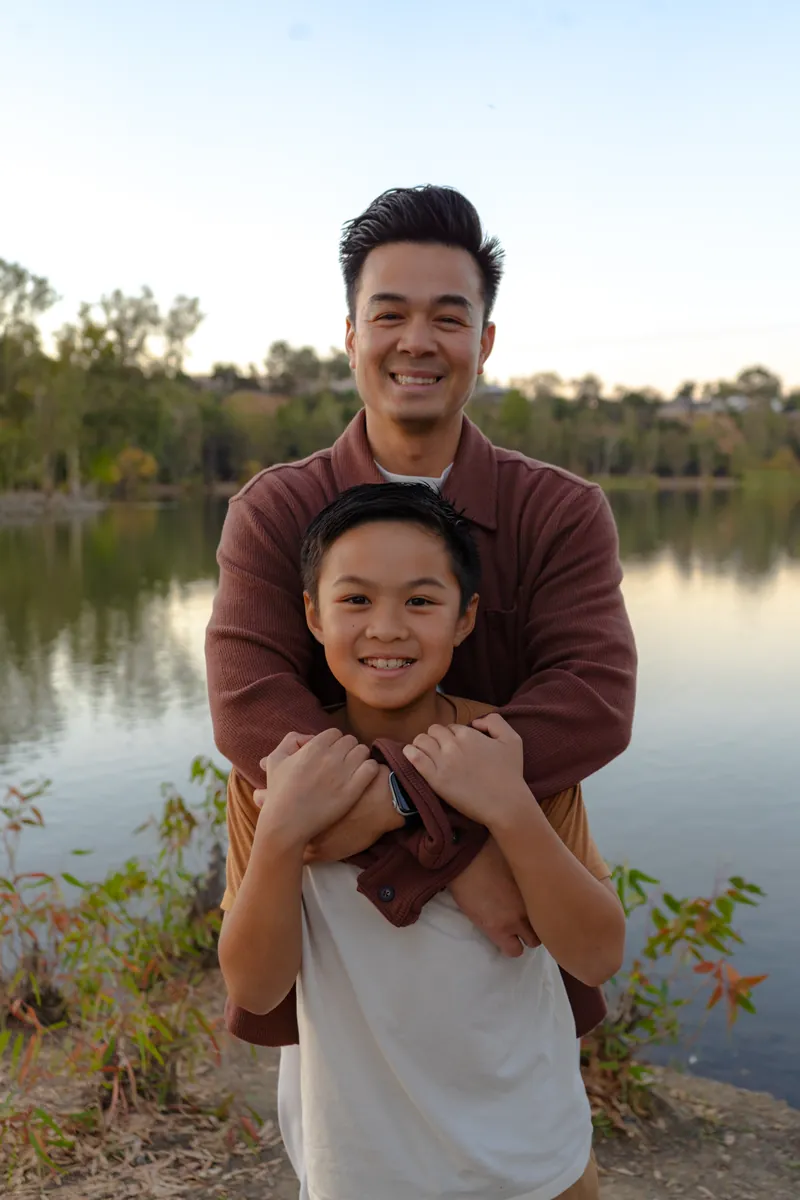 A dad hugging his son from behind by the lake