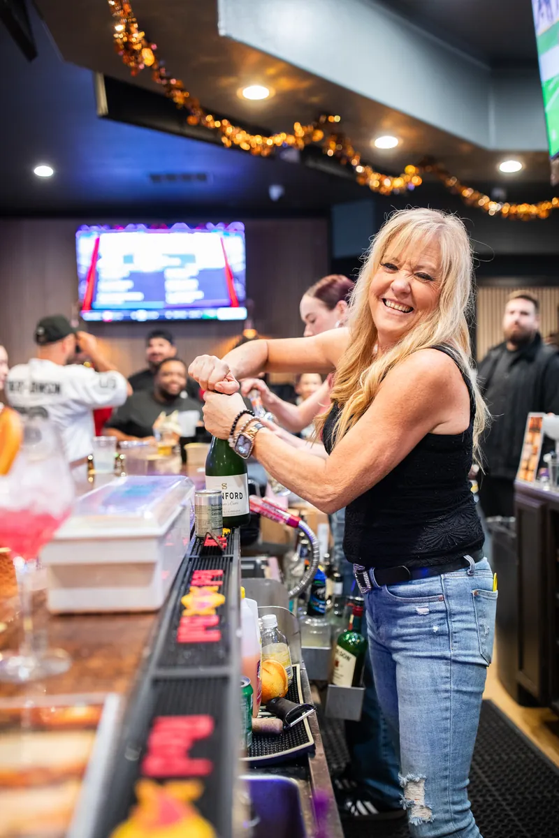 The bartender laughing as she opens a bottle of sparkling wine behind a busy bar