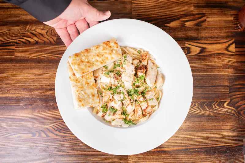 A hand sets down creamy fettuccine with grilled chicken and garlic bread, from above
