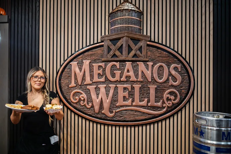 A server holding two plates in front of the Meganos Well sign
