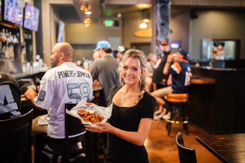 A server holding a basket of wings in the bar, fans in jerseys behind her