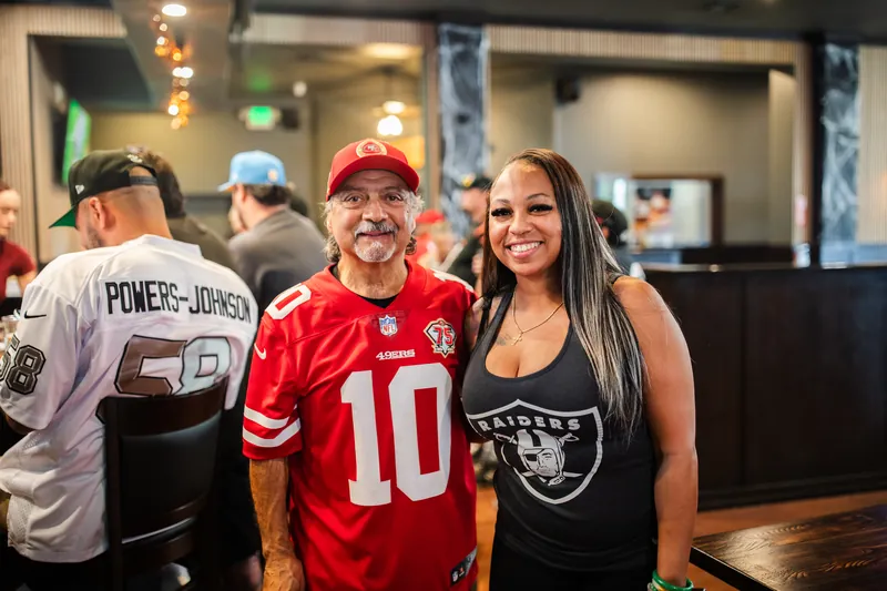 A 49ers fan and a Raiders fan smiling side by side in the bar