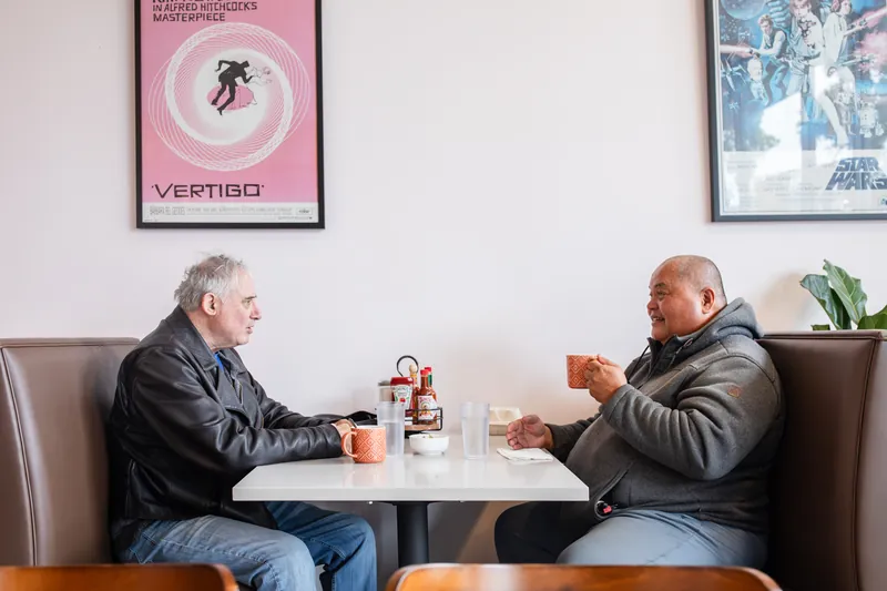 Two regulars talking over coffee in a booth under vintage movie posters